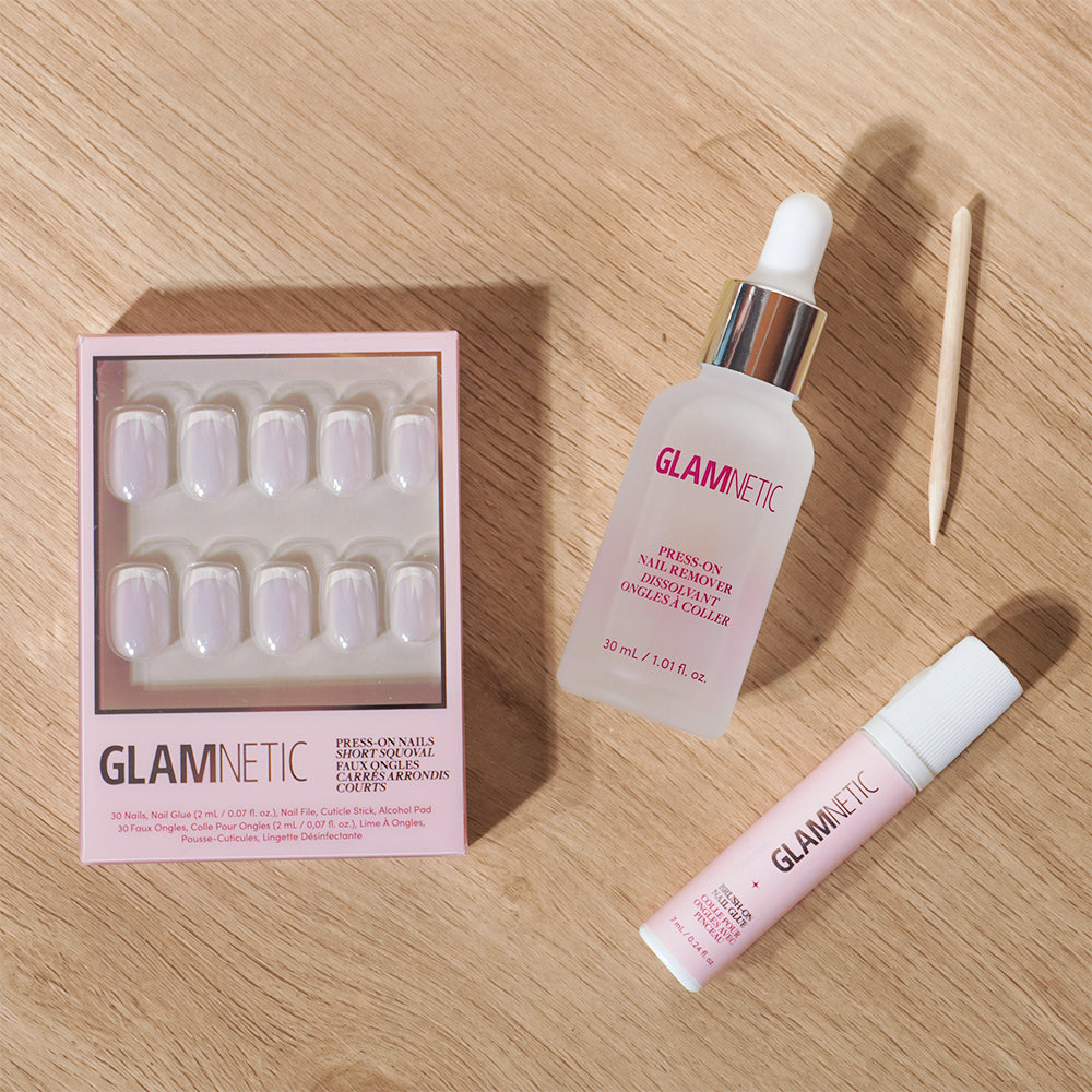A set of glamnetic Press & Perfect press-on nails, along with a dropper bottle of nail remover, a tube of nail glue, and a wooden cuticle stick, are arranged on a light wood surface. Each product features the distinctive “glamnetic” branding. - Product image