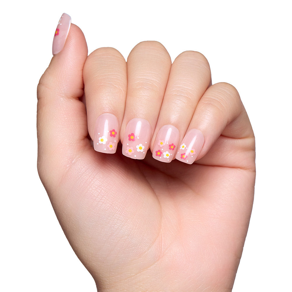 A hand with short squoval nails features glamnetics Pretty Bloom—a glossy light pink polish adorned with small colorful flower designs—beautifully displayed against a white background. - Product image