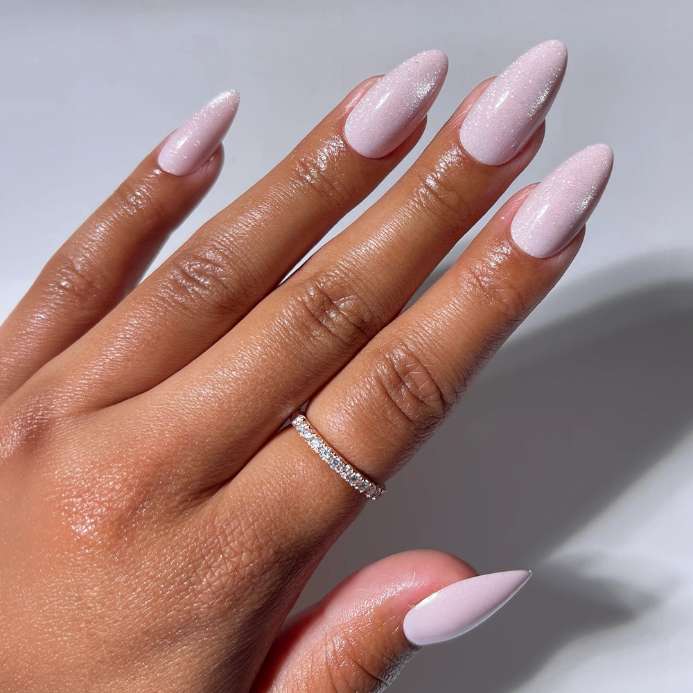 A hand with long, almond-shaped pink nails in "Promise Kept" by glamnetic, featuring a subtle shimmer and a thin silver ring with small stones on the ring finger, is shown against a plain white background.