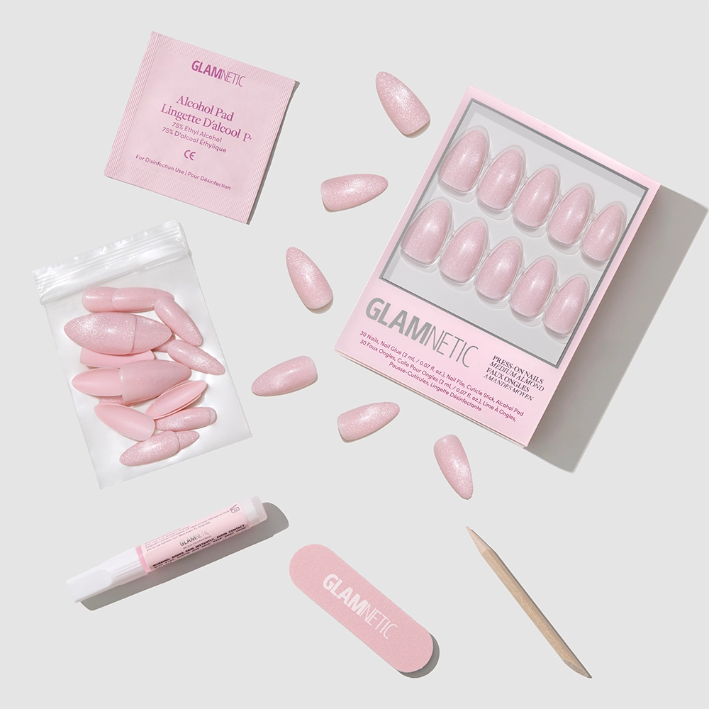 The glamnetic "Promise Kept" kit includes almond-shaped pink press-on nails, a nail file, wooden cuticle stick, nail glue tube, alcohol pad, and extra nails arranged on a light gray surface.