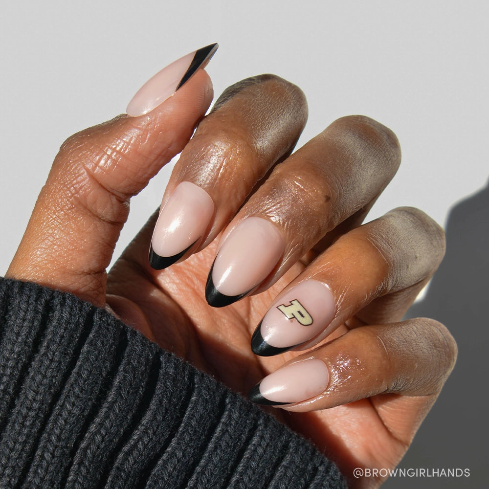 A hand wears the glamnetic Purdue Boilermakers Press-On Nail Set—almond nude nails with black French tips and a gold-and-black P logo accent, ideal for team spirit. The person has on a black ribbed sweater. - Product image