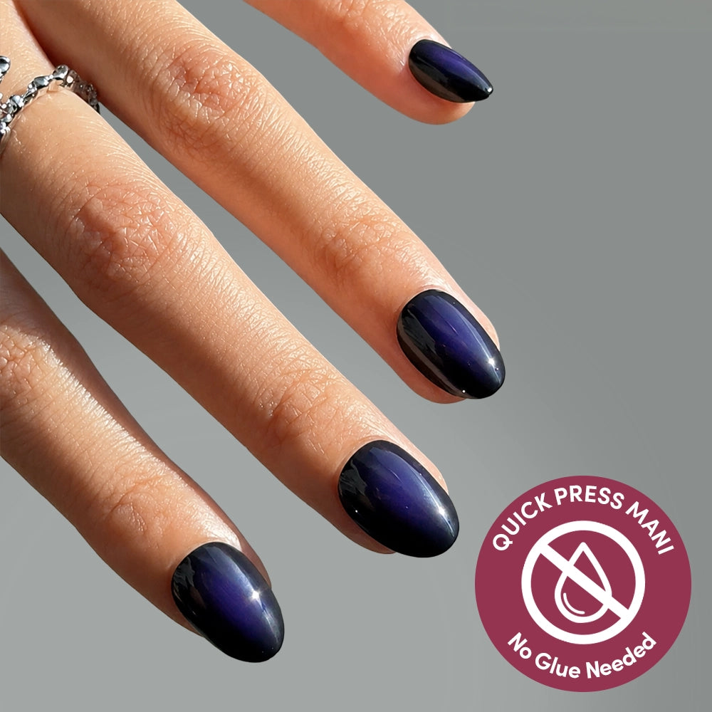 A hand with dark, glossy, almond-shaped Purple Reign press-on nails by glamnetic, adorned with silver rings. A badge reads Quick Press Mani—no-glue nails for effortless style, no adhesive required.