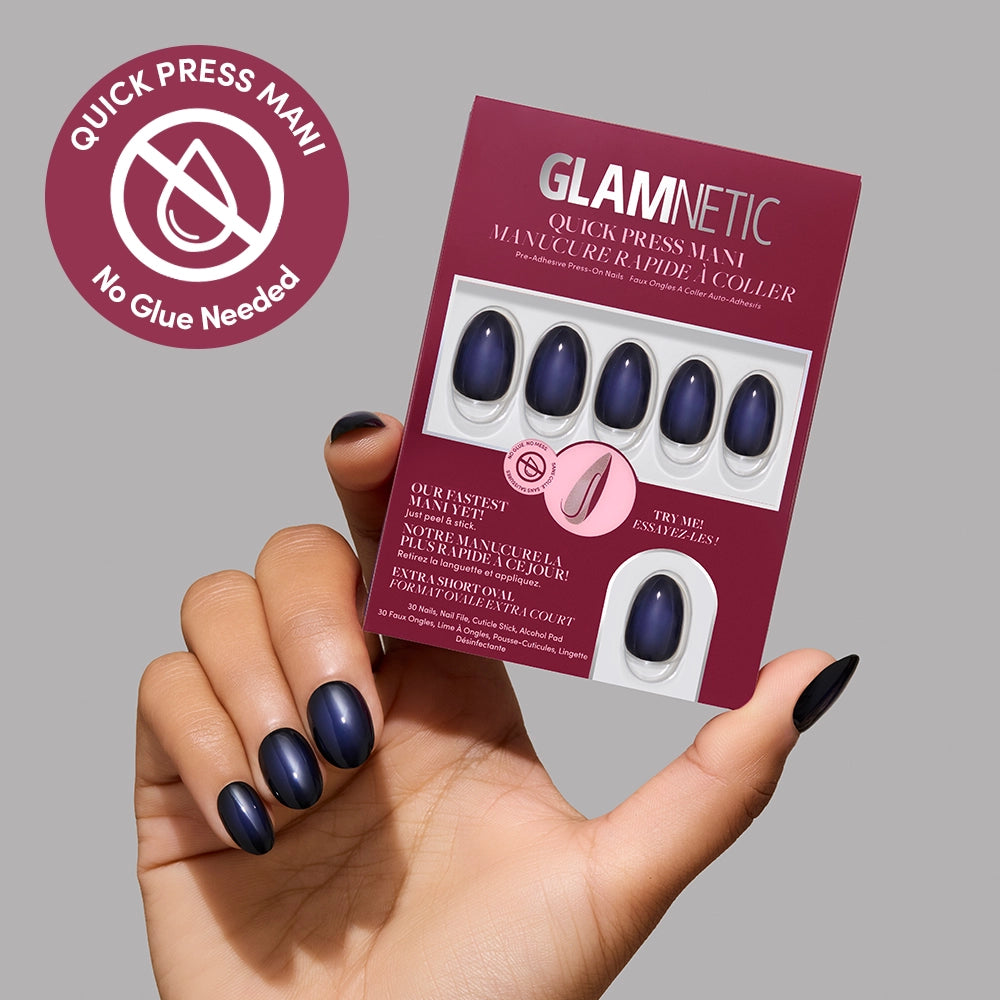 A hand with dark blue manicured nails holds a box of glamnetic Quick Press Mani press-on nails in the shade Purple Reign. The packaging displays a No Glue Needed icon and text in both English and French, emphasizing the convenience of these no-glue stick-on nails. - Product image
