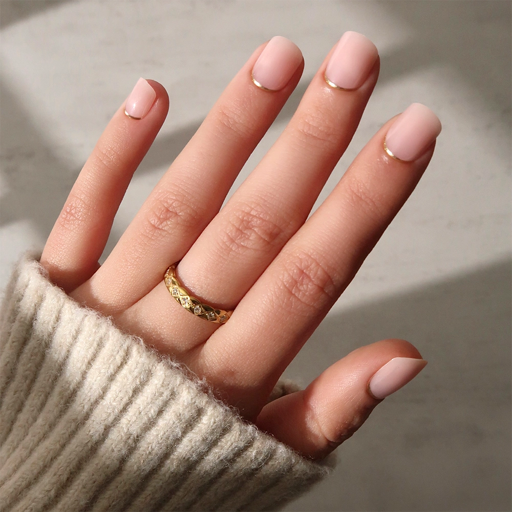 A hand with glamnetic’s Quiet Luxury press-ons—nude nails with gold French tips—wears a gold ring and rests against a neutral background. The person sports a light-colored, ribbed sweater.