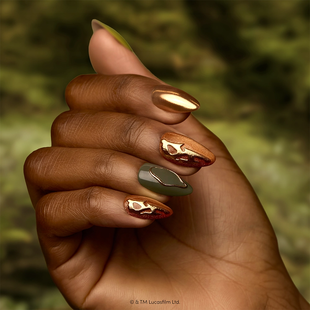 Rebellion - A hand with long, almond-shaped nails—showcasing glamnetic’s Rebellion press-on nails collection—is painted in metallic gold, olive green, and copper with abstract gold designs, set against a blurred green background.
