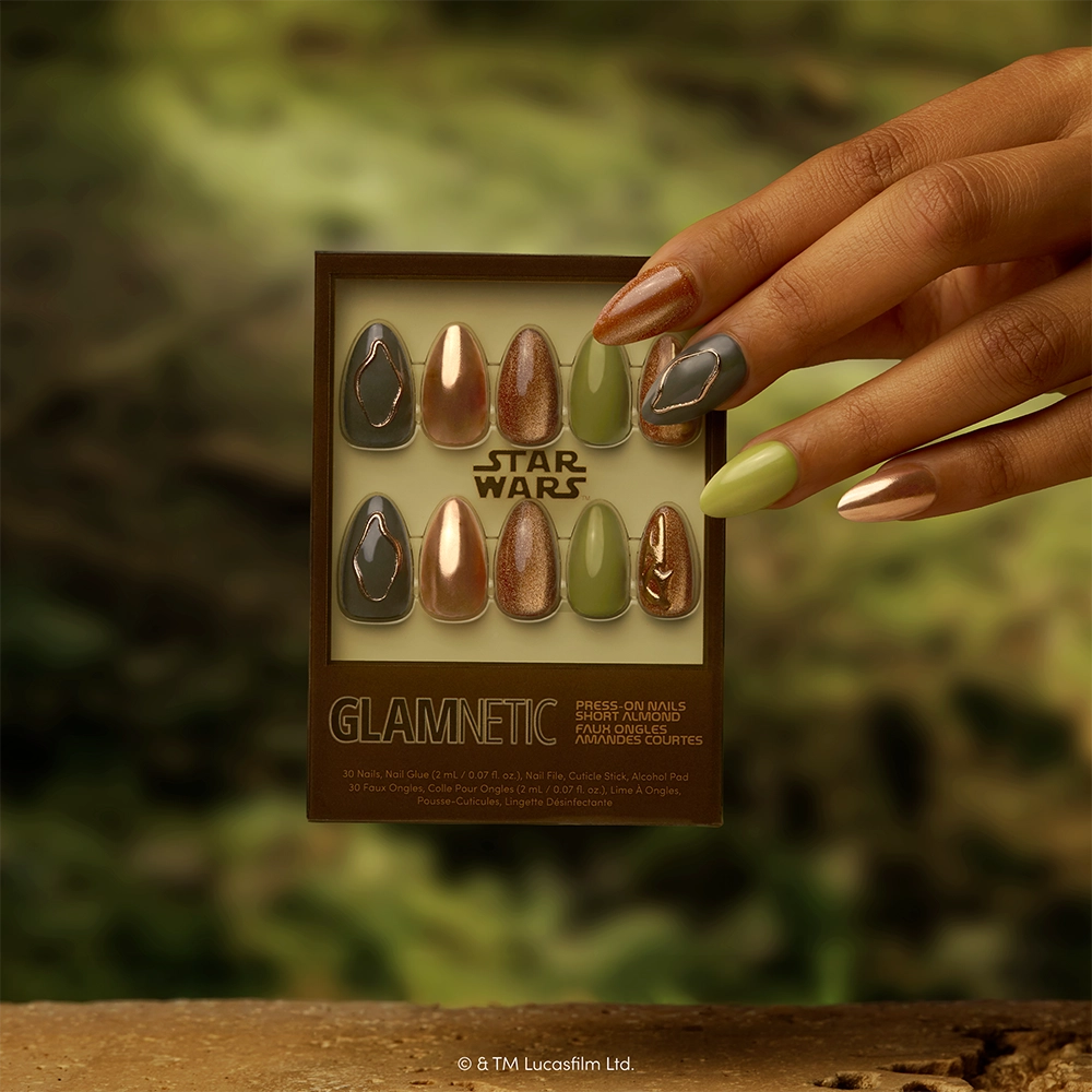 Rebellion - A hand displays a short almond manicure featuring metallic and green STAR WARS press-on nails next to the glamnetic Rebellion Press-On Nail Set—showcasing various designs inspired by Star Wars—against a blurred green nature background.