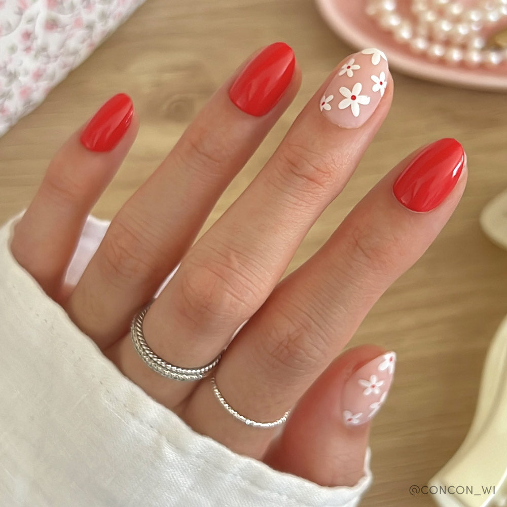 A hand features glamnetics Red Blossom nails—glossy red base, nude tips with white daisies. Silver rings and a white sleeve complete the look. A soft, neutral background with floral accents highlights these chic press-ons.