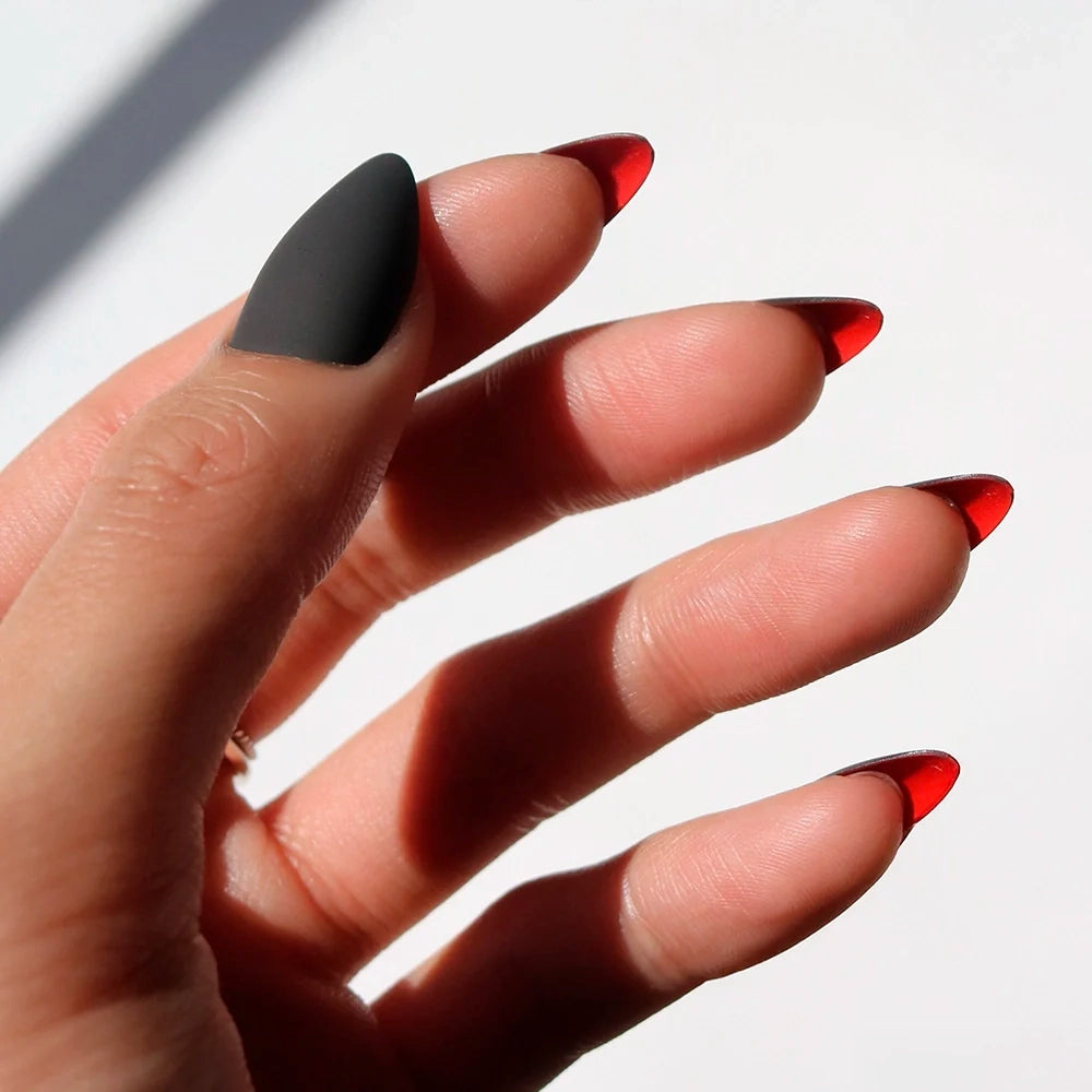 A hand with long Black Matte Almond Nails; the thumb is matte black, and the rest showcase glamnetic Red Bottom nails with glossy red tips in a gradient or flame style.