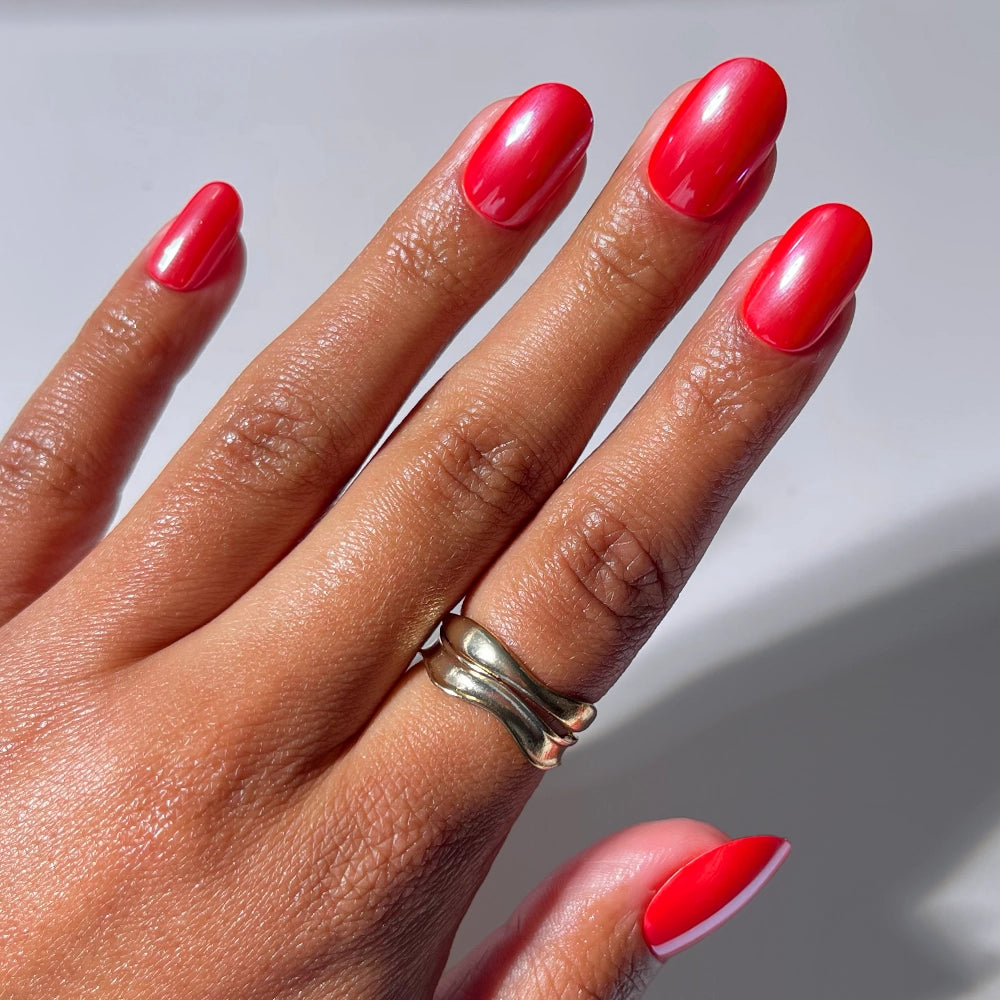 A hand with smooth brown skin models glamnetic's Red Confidence press-on nails in a short round shape, showcasing glossy bright red color. Two gold rings adorn the middle finger against a plain light background for a salon-quality look.