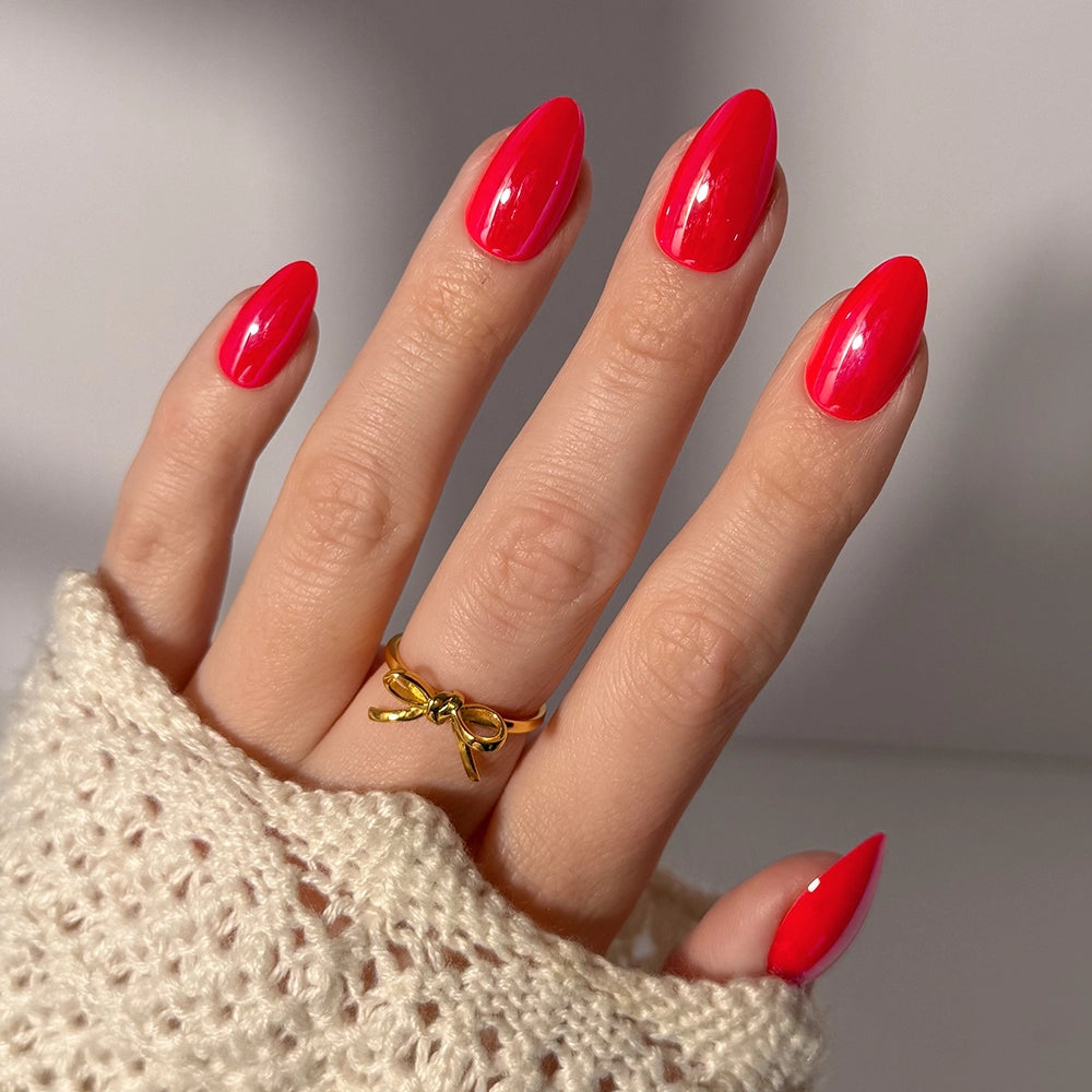 A hand with long, glazed red nails wears a gold bow ring. Styled in a cream knit sleeve, the elegant vegan, cruelty-free "Red Flag" press-on nails by glamnetic are softly lit in the background.