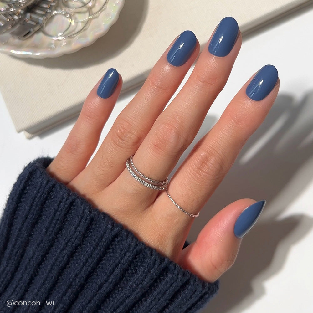 A hand with neatly manicured nails painted in Regal Blue matte polish, wearing two delicate silver rings and a navy ribbed sweater. The scene includes a white surface and some jewelry in the background. - Product image