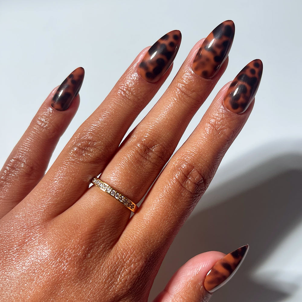 A hand with almond-shaped glossy brown and black tortoiseshell “Rich Behavior” press-on nails by glamnetic is shown. The person wears a gold ring with clear stones, highlighting the salon-quality manicure. - Product image