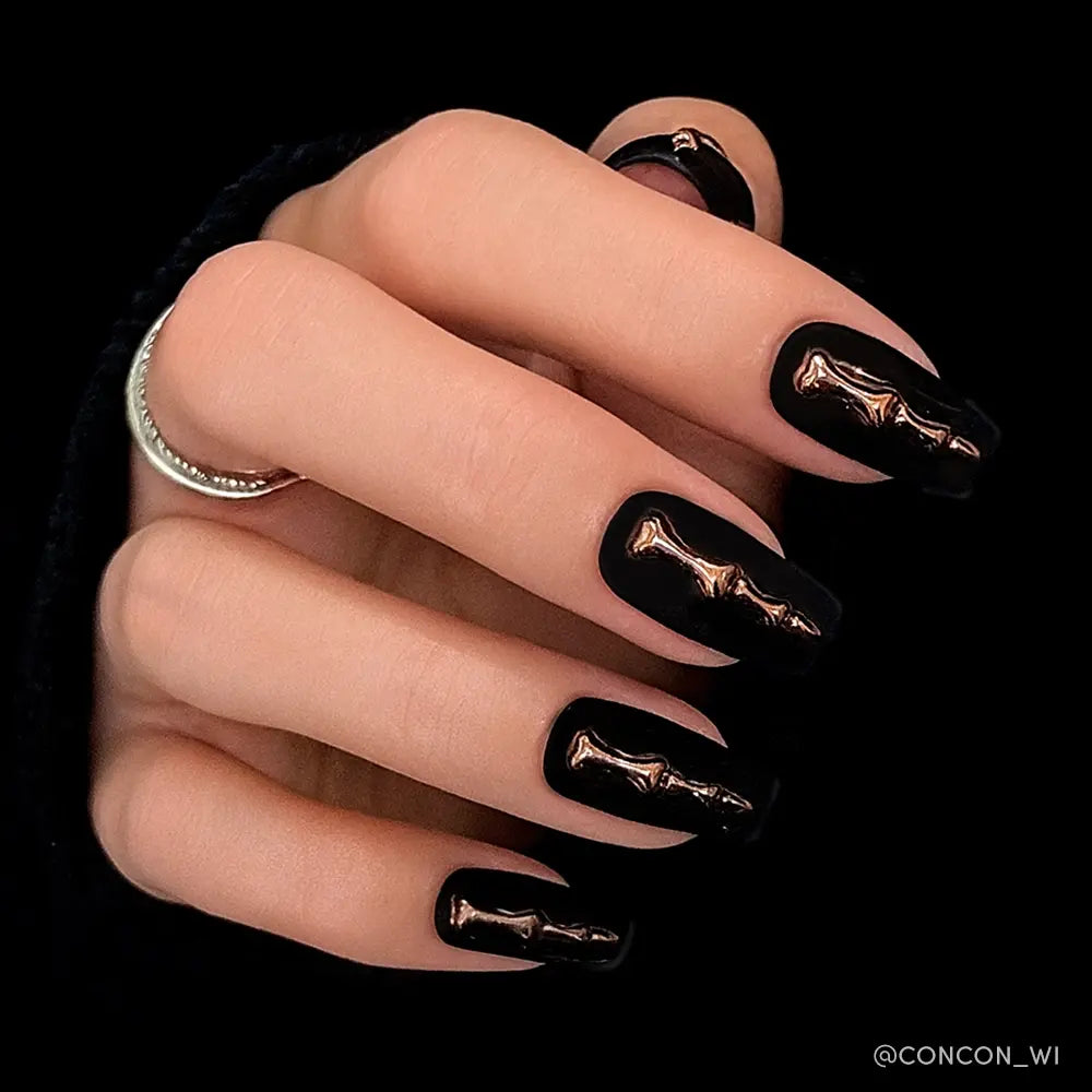 A hand shows off glamnetic’s Rosy Remains: matte charcoal coffin nails with shiny, metallic 3D bone art. The person wears a silver ring and a black sleeve. - Product image