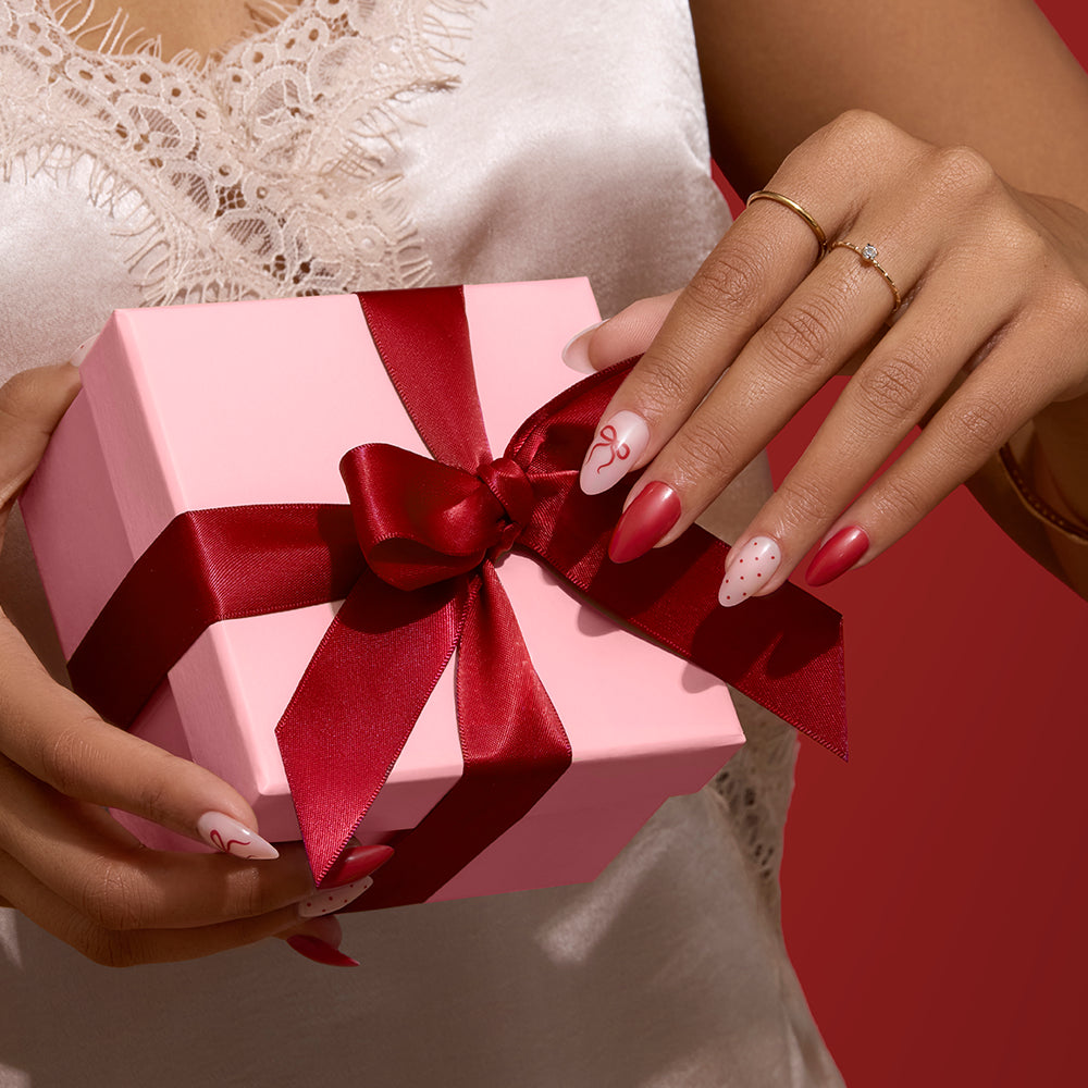 Royal Ribbons - Hands holding a pink gift box tied with a dark red satin ribbon, showcasing a Glamnetic Royal Ribbons holiday manicure with almond nails, red bows, and polka dot designs.
