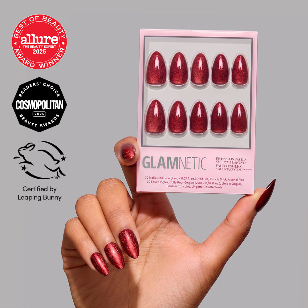 Ruby Slipper - A hand holding a Glamnetic Ruby Slipper nail set of short almond-shaped red glitter press-on nails, featuring Allure and Cosmopolitan award seals and a Leaping Bunny certification logo.