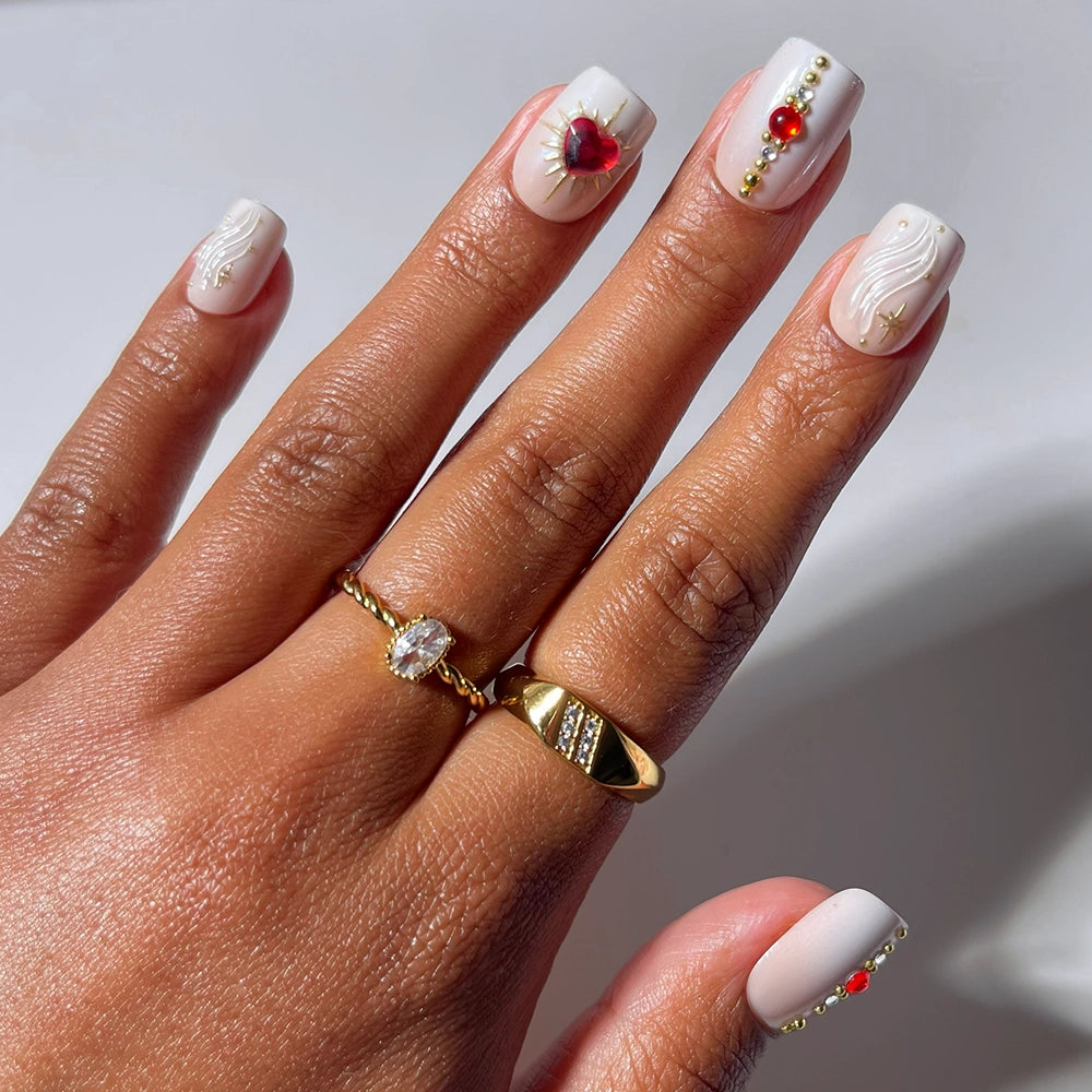A hand showcases light pink, salon-quality press-on nails in a square shape from glamnetic's Ruby collection, adorned with gold and red gem accents. The look is complemented by two gold rings—one featuring a clear stone and the other with a textured design—perfectly enhancing the ruby-inspired elegance of Ruby by glamnetic.