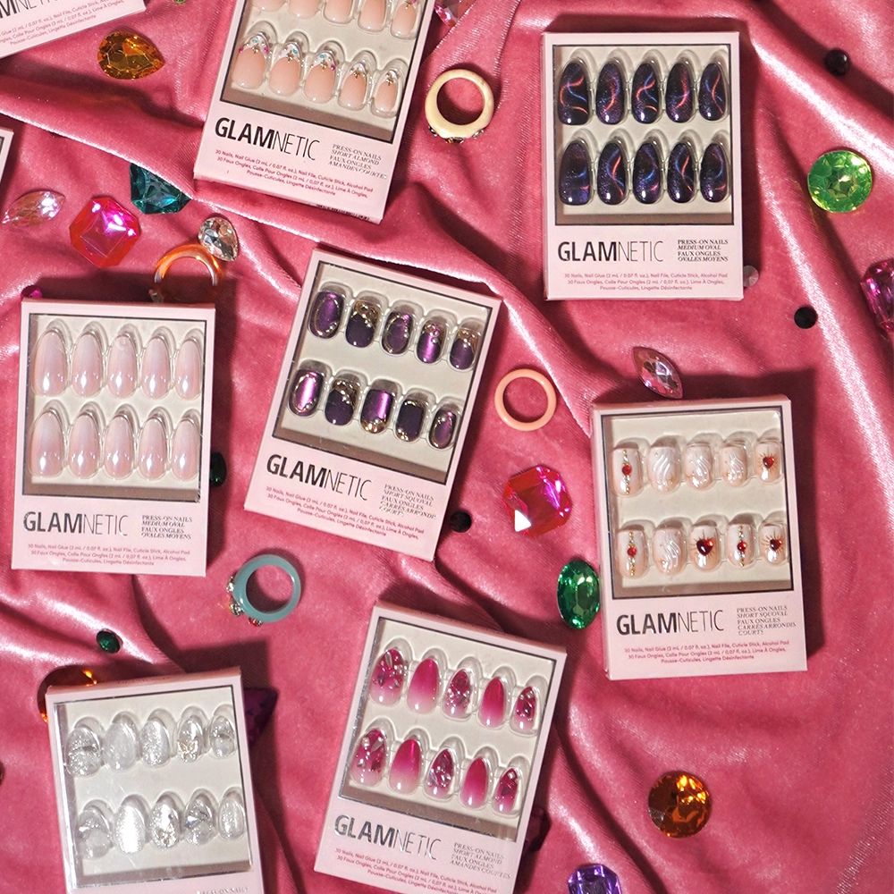Boxes of glamnetic Ruby press-on nails, featuring a variety of salon-quality nail art designs, are displayed on pink satin fabric and surrounded by colorful plastic gems, rings, and decorative accents. - Product image