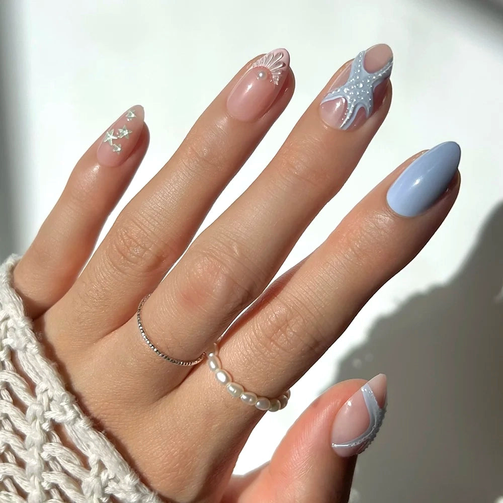 A hand showcases “Salty Waves” by glamnetic, featuring light blue and nude sea-inspired nail art with pearls, white lines, and delicate dots on short almond nails. Two rings adorn the fingers, complemented by a crocheted sleeve.