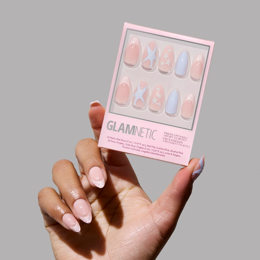A hand with manicured short almond nails holds a box of glamnetic Salty Waves Press-On Nail Set. The pastel pink and blue nails feature stars, swirls, and solids for a chic ocean-inspired look against a plain gray background.
