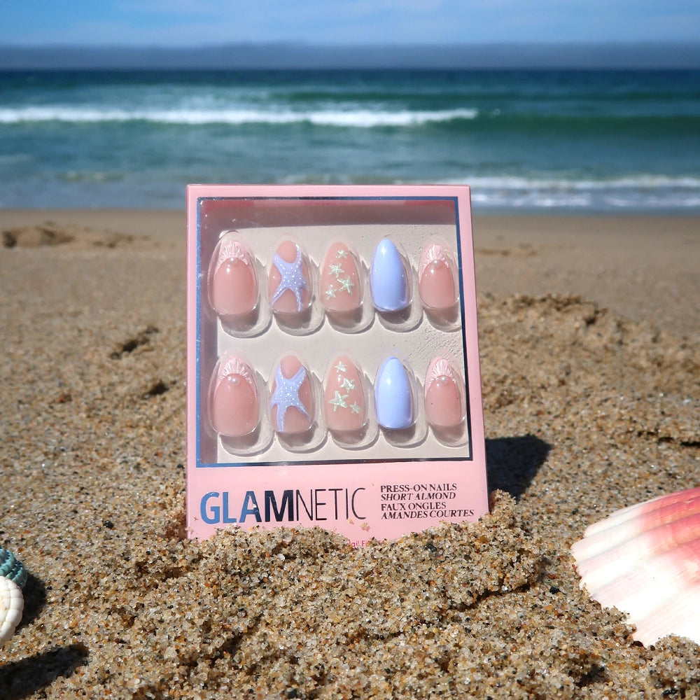 A box of glamnetic Salty Waves Press-On Nail Set, featuring pastel pink and blue ocean-inspired nail art with starfish designs, rests on the sandy beach among seashells with the ocean and a clear sky in the background. - Product image