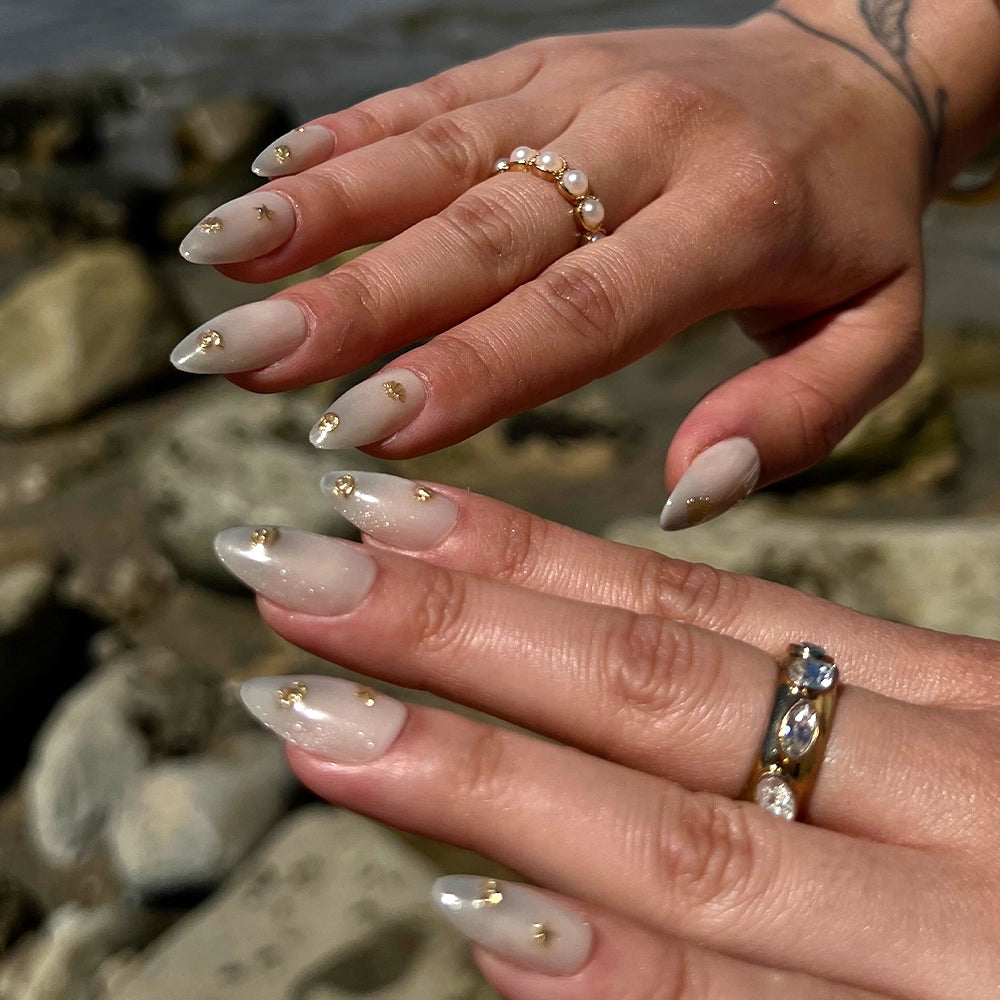Two hands with long, almond-shaped Sanded nails by glamnetic feature a soft ombre design and gold details for subtle mermaid vibes. Pearl and gemstone rings enhance the beachy look against a blurred rocky background. - Product image