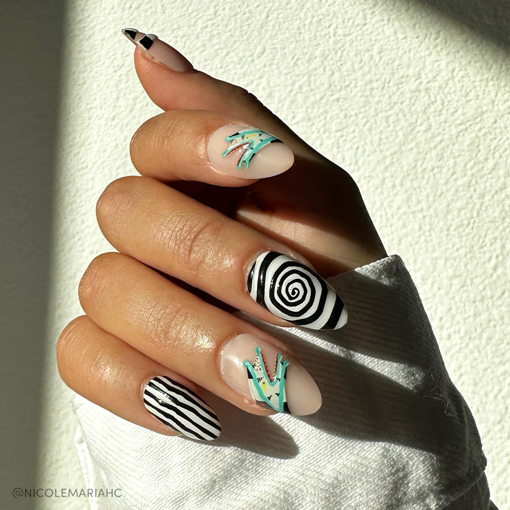A hand with almond-shaped nails showcases abstract nail art inspired by glamnetics Sandworm set, with black and white stripes, a black spiral, teal and white accents, and a black triangle on nude. A white sleeve partially covers the hand.