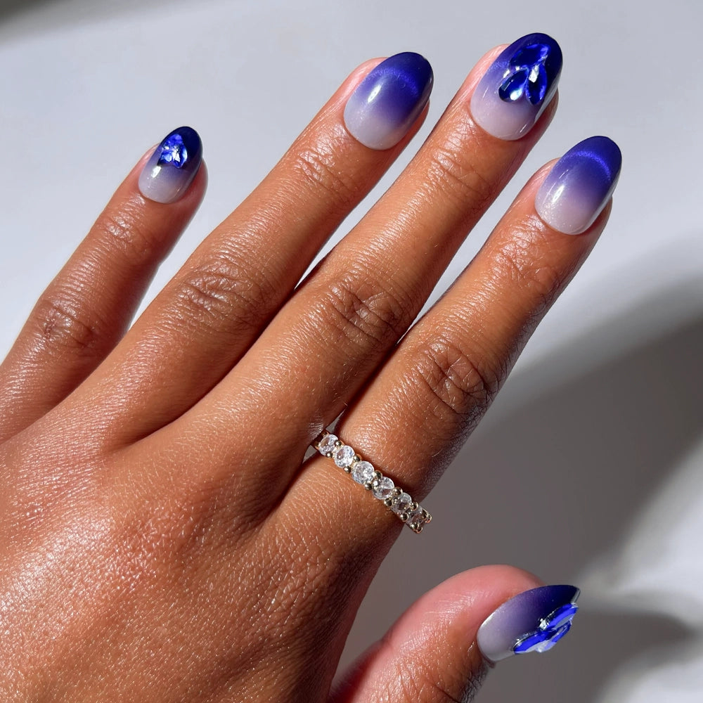 A hand adorned with a gold and diamond ring displays short, oval-shaped nails featuring glamnetic’s Sapphire Press-On Nails—a blue-to-clear ombré design embellished with delicate blue floral accents and sparkling 3D sapphire-inspired gemstones.