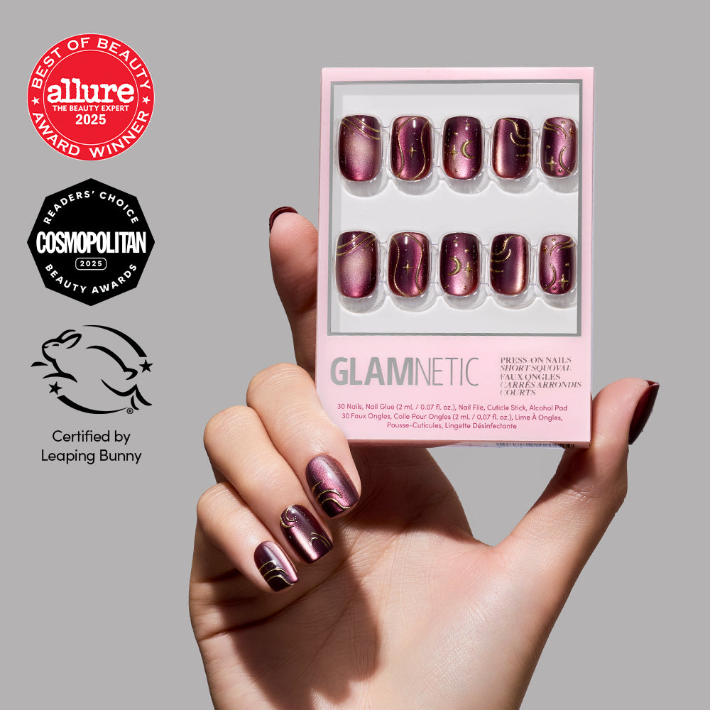 A hand with glossy maroon and gold marble-patterned Scarlet Spell press-on nails by glamnetic, styled in a short squoval shape reminiscent of red-brown velvet nails, holds a box of glamnetic nails; three award badges and a leaping bunny certification logo are displayed on a grey background. - Product image