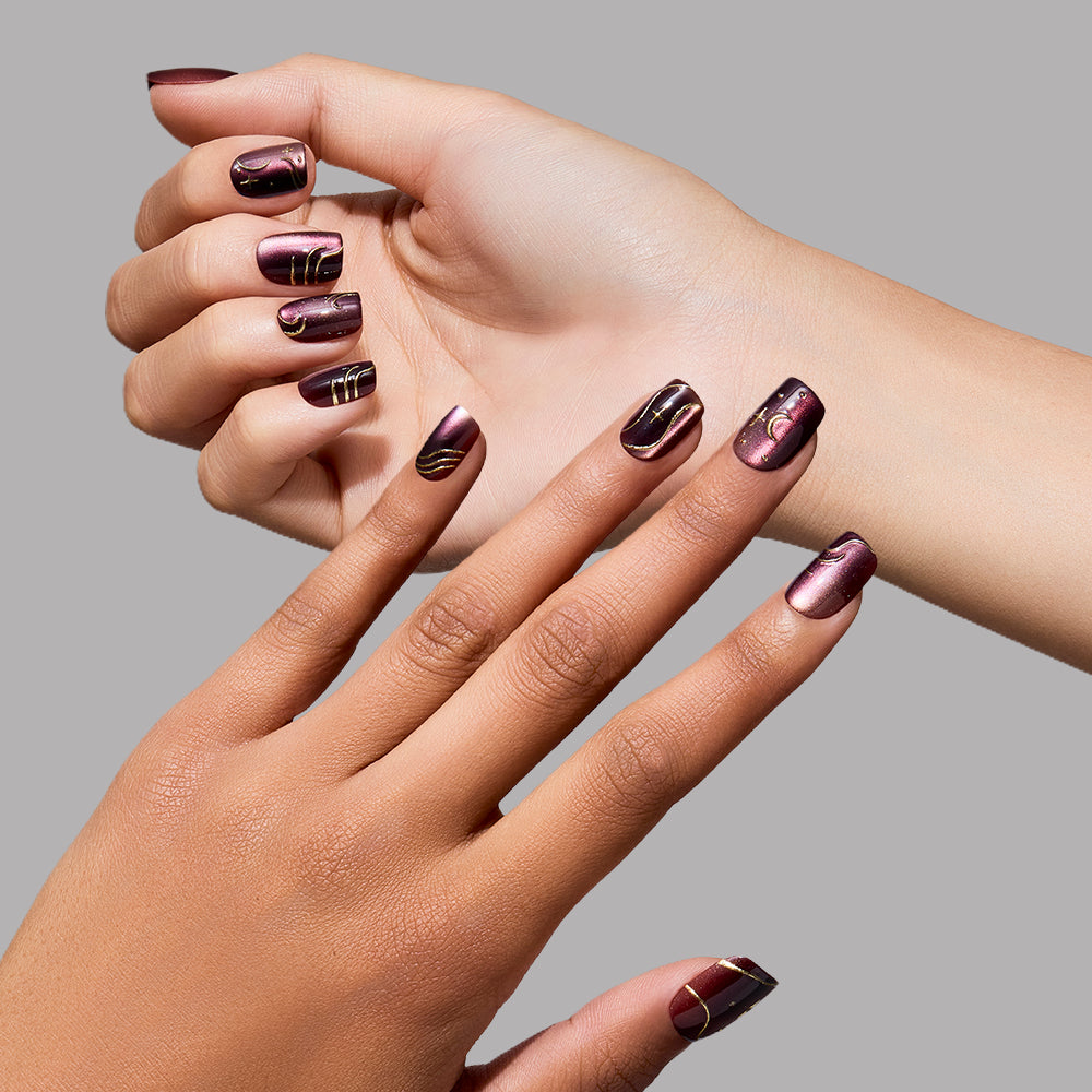 Two hands with short squoval nails painted in glamnetic’s Scarlet Spell—a dark, glossy maroon shade—are shown against a plain gray background, featuring gold and black abstract line designs. - Product image