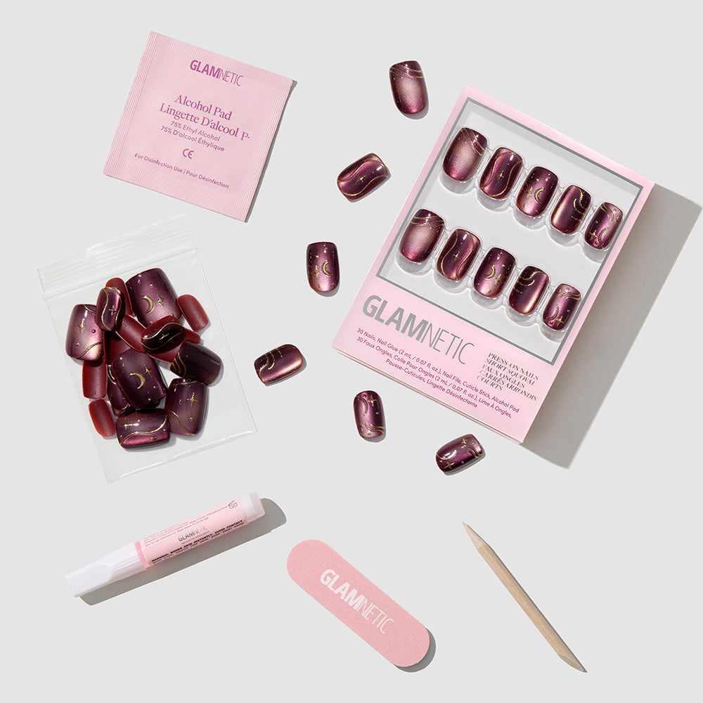 The Scarlet Spell press-on nails by glamnetic, along with nail glue, a nail file, a cuticle stick, an alcohol pad, and extra nails, are arranged neatly on a light gray background. These short squoval pink metallic nails deliver an effortlessly chic look for any occasion. - Product image