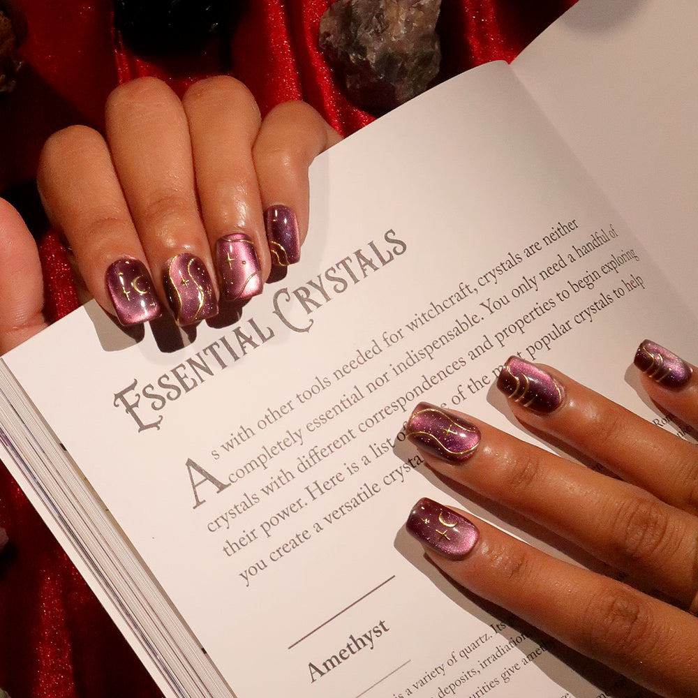 A hand with shiny purple, short squoval nails painted in Scarlet Spell by glamnetic holds open a book titled Essential Crystals on a red surface, with crystals placed around the book. - Product image