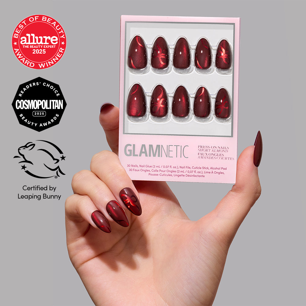 A hand holds a box of Glamnetic Scarlet Ribbons short almond velvet press-on nails featuring a deep red ribbon design, alongside Allure and Cosmopolitan award logos and a Leaping Bunny certification.