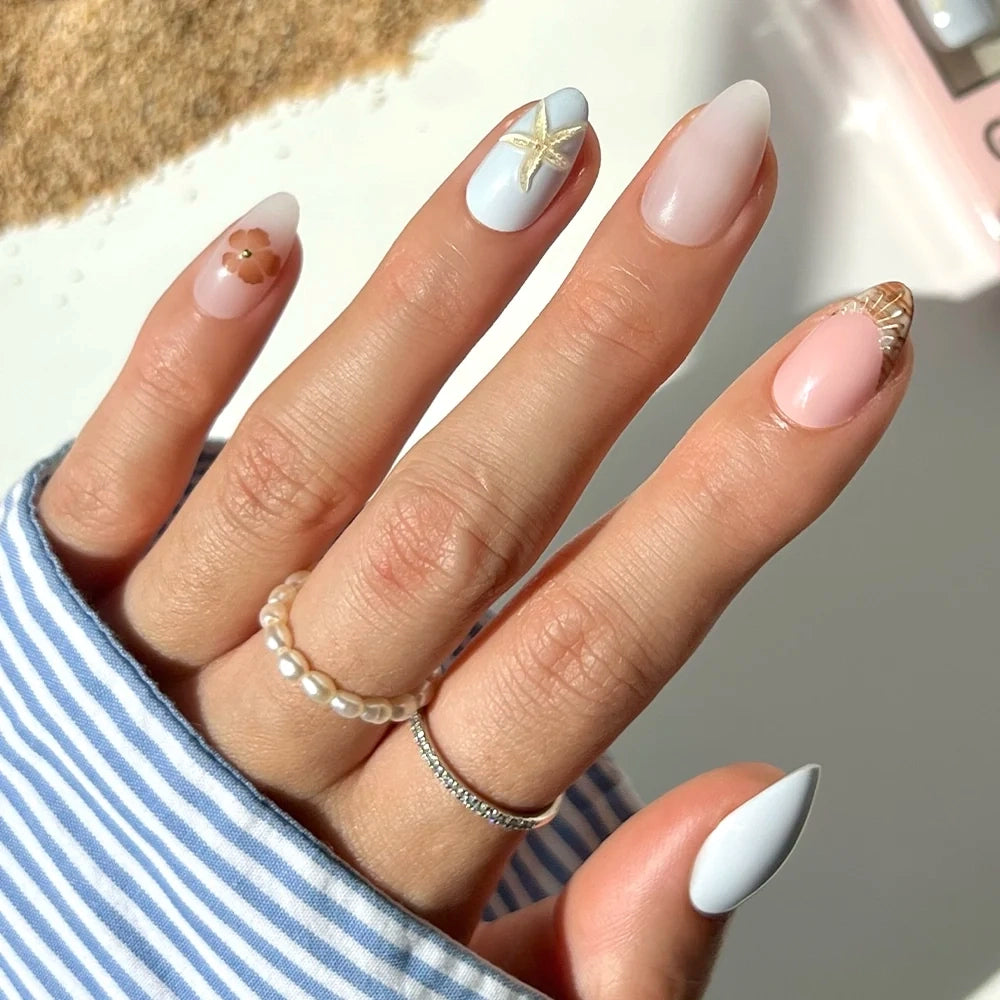 A hand showcases long, almond-shaped nails adorned with glamnetic’s Sea Star set—pastel nail art featuring a starfish, shells, and delicate gold accents. The subtle seashell designs bring a coastal charm to the look. The person complements the manicure with pearl and silver rings and a blue-striped shirt.