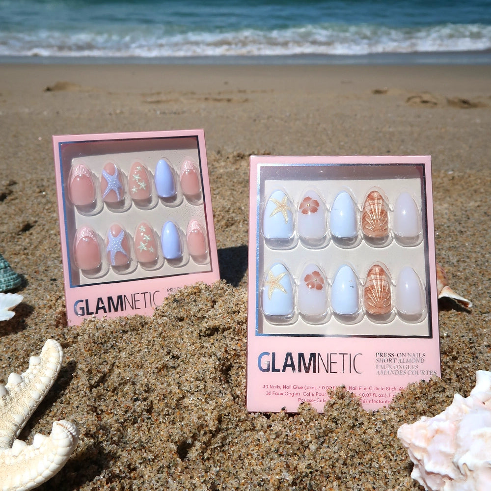 Two boxes of glamnetic Sea Star press-on nails with beachy 3D shell accents stand upright in the sand, surrounded by seashells and ocean waves in the background. - Product image