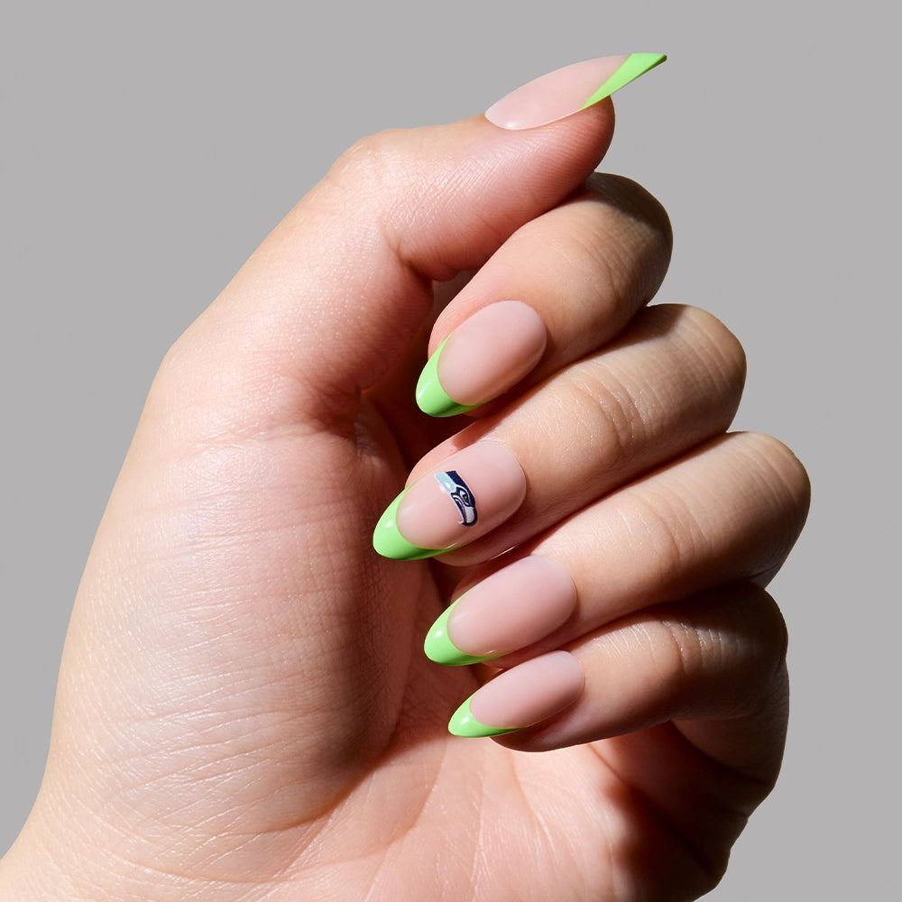 A hand wears glamnetics Seattle Seahawks Press-On Nail Set: almond nude nails with bright green tips and a blue-and-white accent on the ring finger—perfect for subtle fan style—all shown against a plain gray background. - Product image