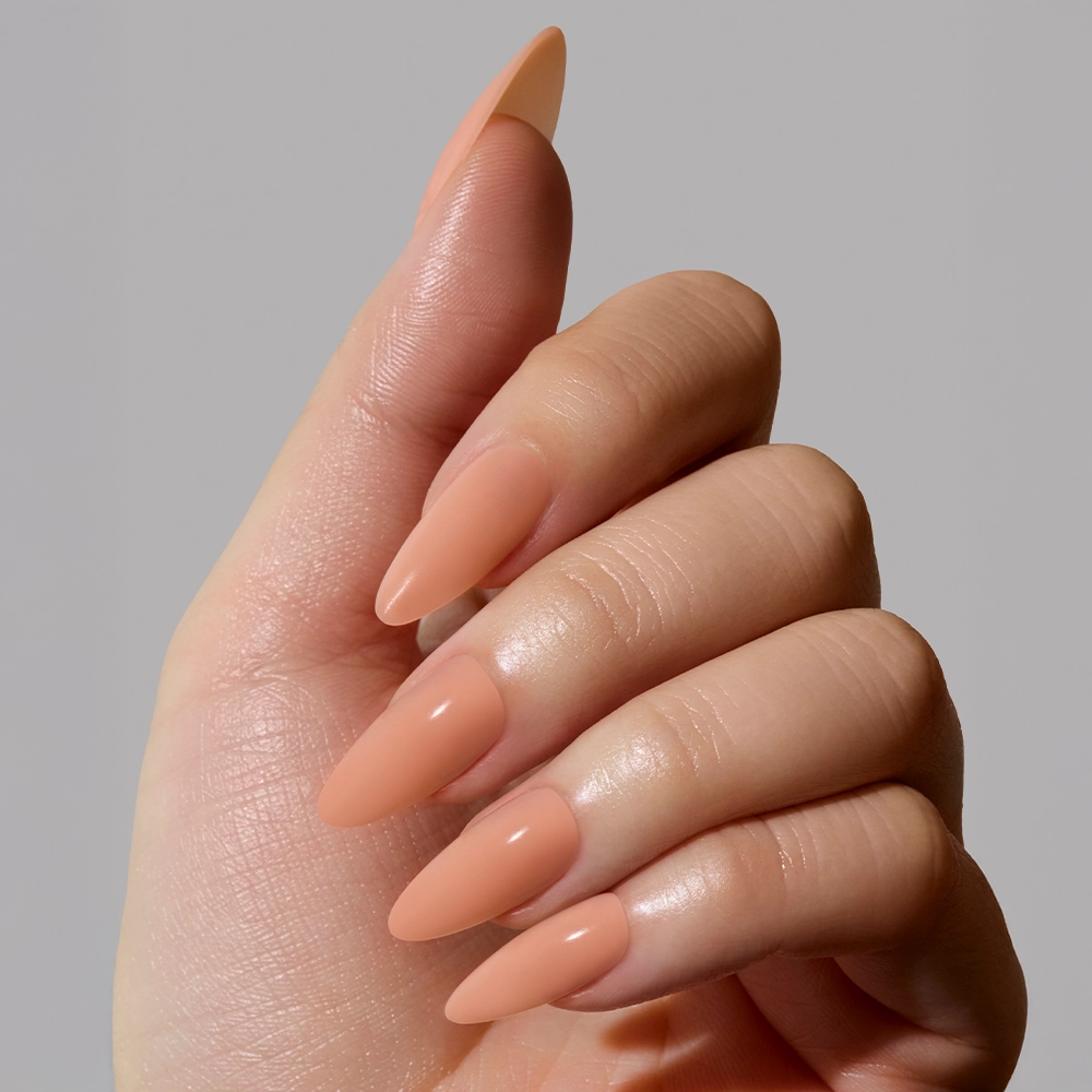 A hand with long, almond-shaped nails painted in glamnetics Sheer Peach, a smooth matte nude shade, is shown against a plain light gray background. - Product image