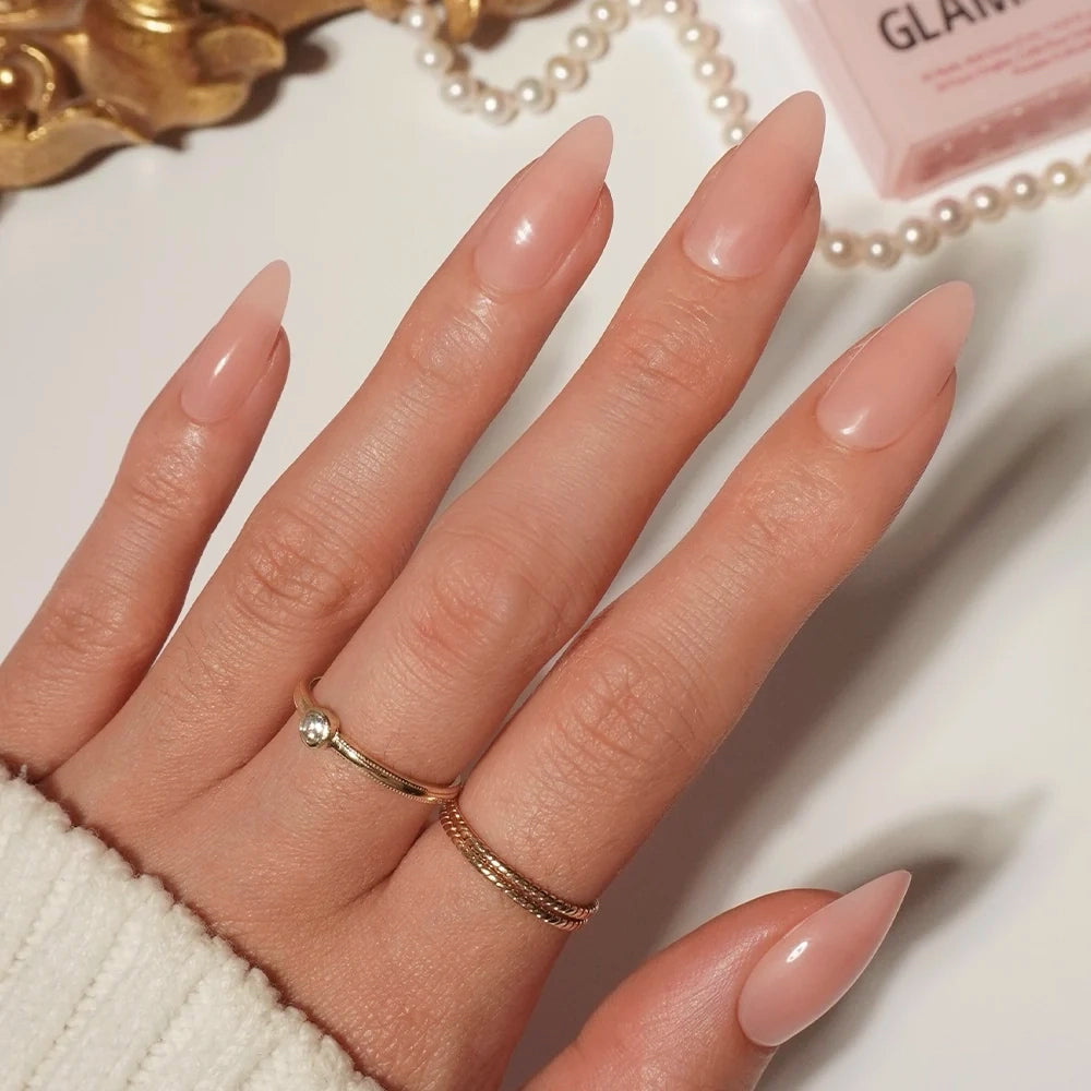 A hand adorned with glamnetic Sheer Pink press-on nails in a long almond shape, accented by two gold rings, rests on a white surface next to scattered pearls and a pink box—perfectly capturing the clean-girl aesthetic.