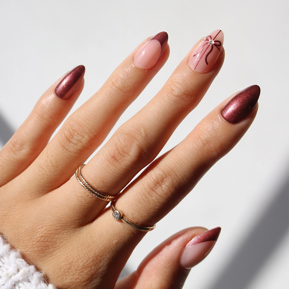 A hand showcases glamnetic’s “She’s A Gift” press-on nails, featuring almond shapes painted in metallic burgundy and nude shades. One nail is adorned with a bow design and accented by a small rhinestone. The look is completed with two gold rings on the person’s fingers.