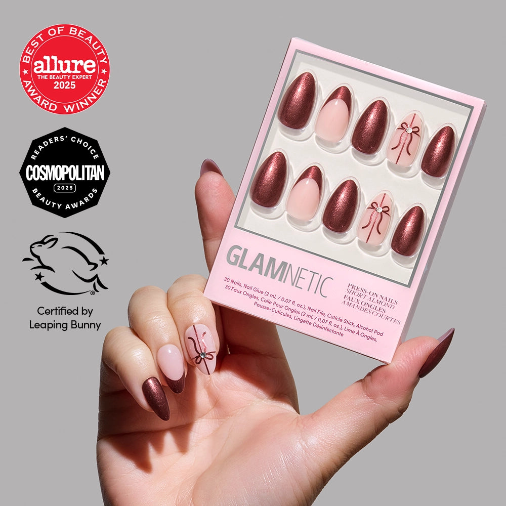 A hand with metallic burgundy press-on nails holds a glamnetic She’s A Gift nail set. The set features shiny burgundy and pink almond-shaped nails embellished with bow designs, ideal for anyone who loves a chic nude manicure. Award and certification logos appear on the left side.