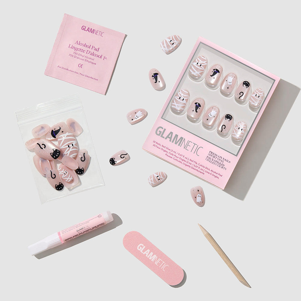 Introducing Shriek Boutique by glamnetic: a set of pink press-on nails with playful cat and mummy designs in a short oval shape. This mani kit comes with nail glue, a pink nail file, a wooden cuticle stick, an alcohol pad, and extra nails—all beautifully arranged on a light gray background. - Product image