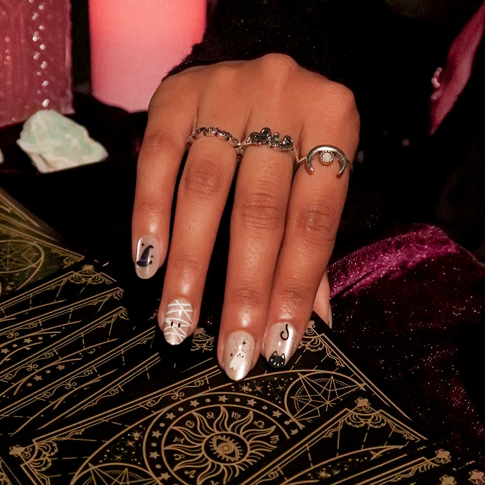 A person’s hand showcases glamnetic’s Shriek Boutique press-on nails—a Halloween-themed set adorned with witch hats, mummy wraps, a ghost, and a black cat—resting on a gold-embossed tarot card. Their short oval mani is perfectly suited for the look, accented by three silver rings (including one with a crescent moon) against a dark background, giving this glamnetic set an enchanting touch of magic. - Product image