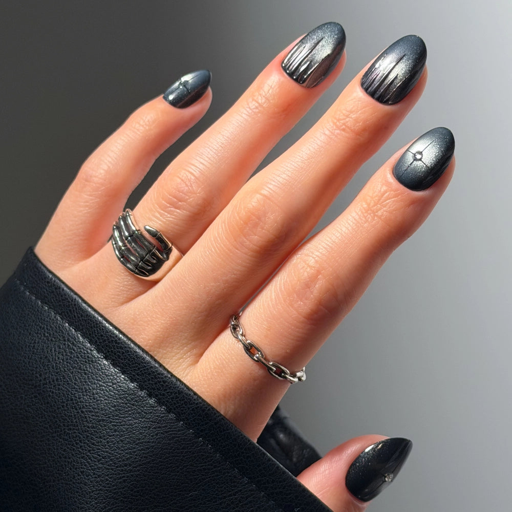 A hand adorned with gradient silver and black metallic nails from glamnetic’s Silver Daggers Press-On Nail Set wears two silver rings against a light background. The black leather sleeves highlight the bold, jewelry-inspired nails.
