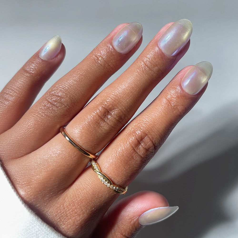 A hand with neatly manicured, almond-shaped nails painted in glamnetic's Silver Siren iridescent blue chrome polish. Two gold rings accent the medium brown skin tone against a softly blurred background.