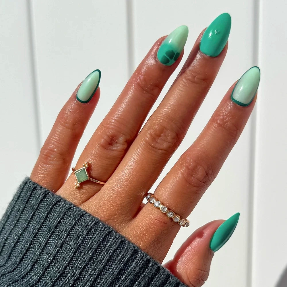 A hand adorned with glamnetic’s Siren Green press-on nails—long, almond-shaped with an aquamarine-inspired green gradient and dark green French tips, plus unique accent designs—is showcased wearing two rings against a white background.