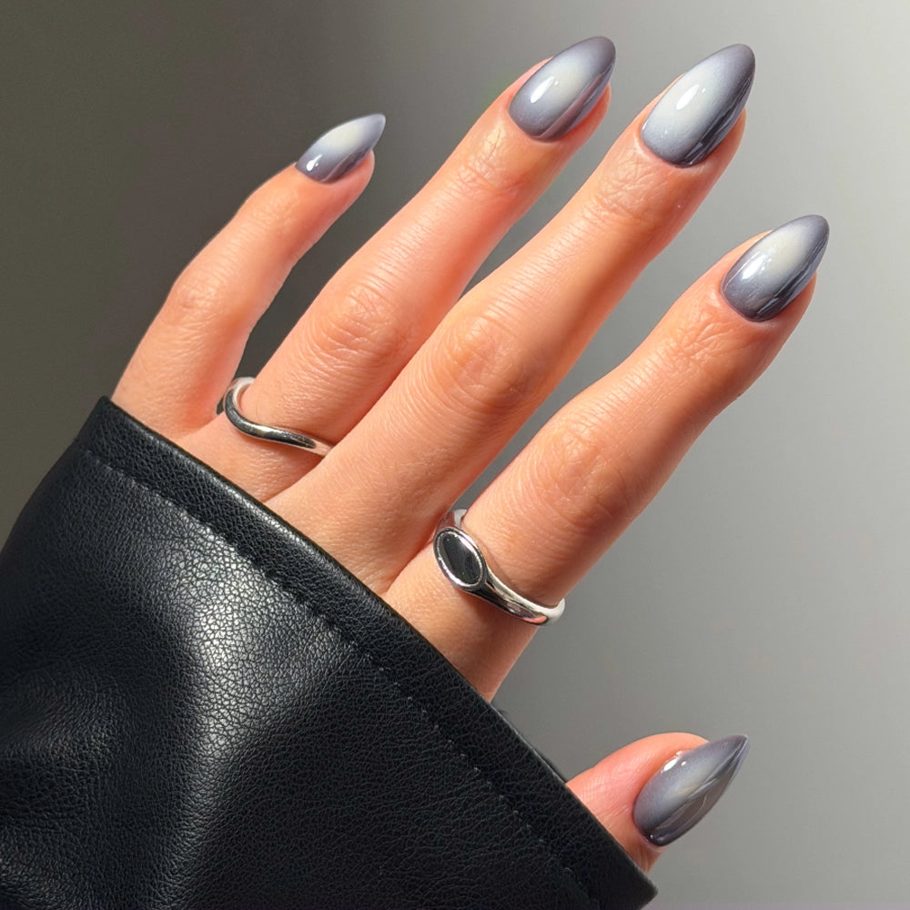 A hand adorned with glamnetic’s Smoke N’ Mirrors press-ons—long, almond-shaped, and featuring a glossy gray finish for salon-quality durability—wears two silver rings and a black sleeve partially covering the wrist.
