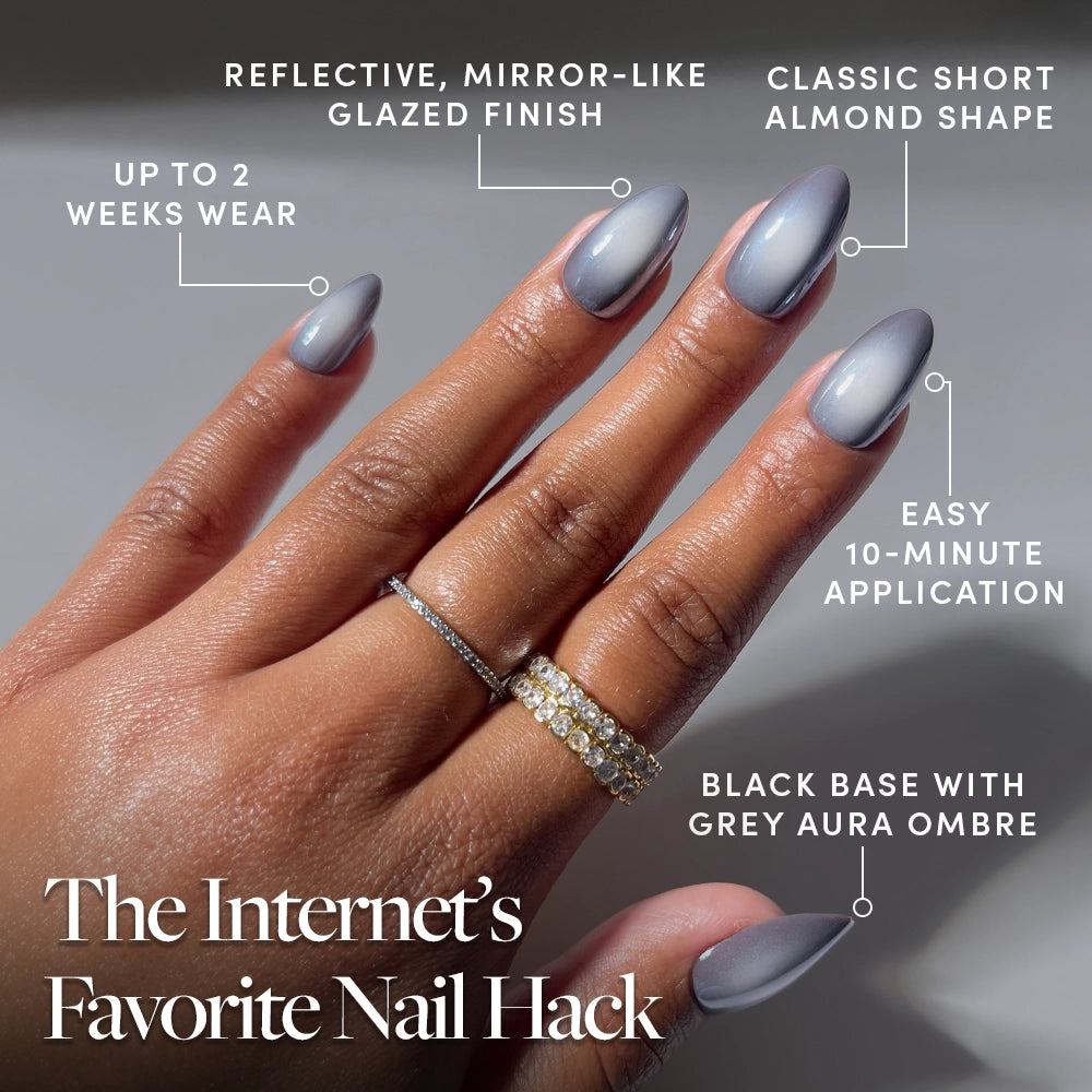 A hand with short almond nails painted in glamnetic’s “Smoke N’ Mirrors,” a reflective grey ombré press-on, sports two rings. Text highlights the fast application, salon-quality durability, long wear, and mirror-like glaze of these press-ons. - Product image