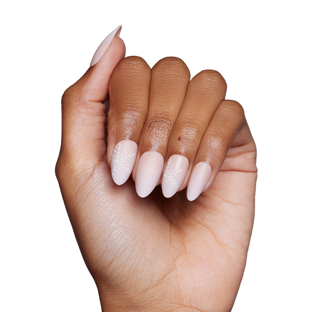 A hand showcases glamnetics Snowdrift nails in a matte nude gradient almond style with delicate snowflake accents on the middle and ring fingers, set against a plain white background. - Product image