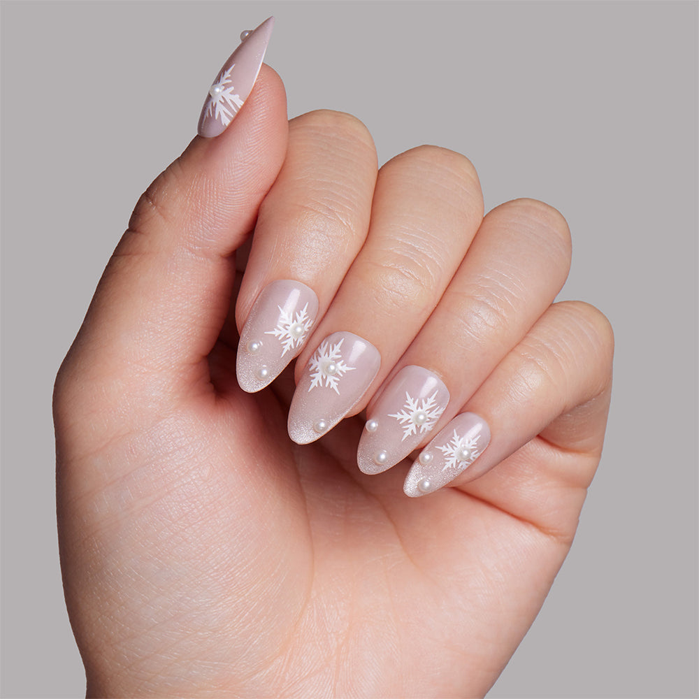 A hand with long, almond-shaped nails painted pale pink, featuring white snowflake designs and small white dots on each nail. The Snowfall Press-On Nail Set from glamnetic delivers a salon-quality finish, making it easy to achieve this look against a simple light gray background. - Product image