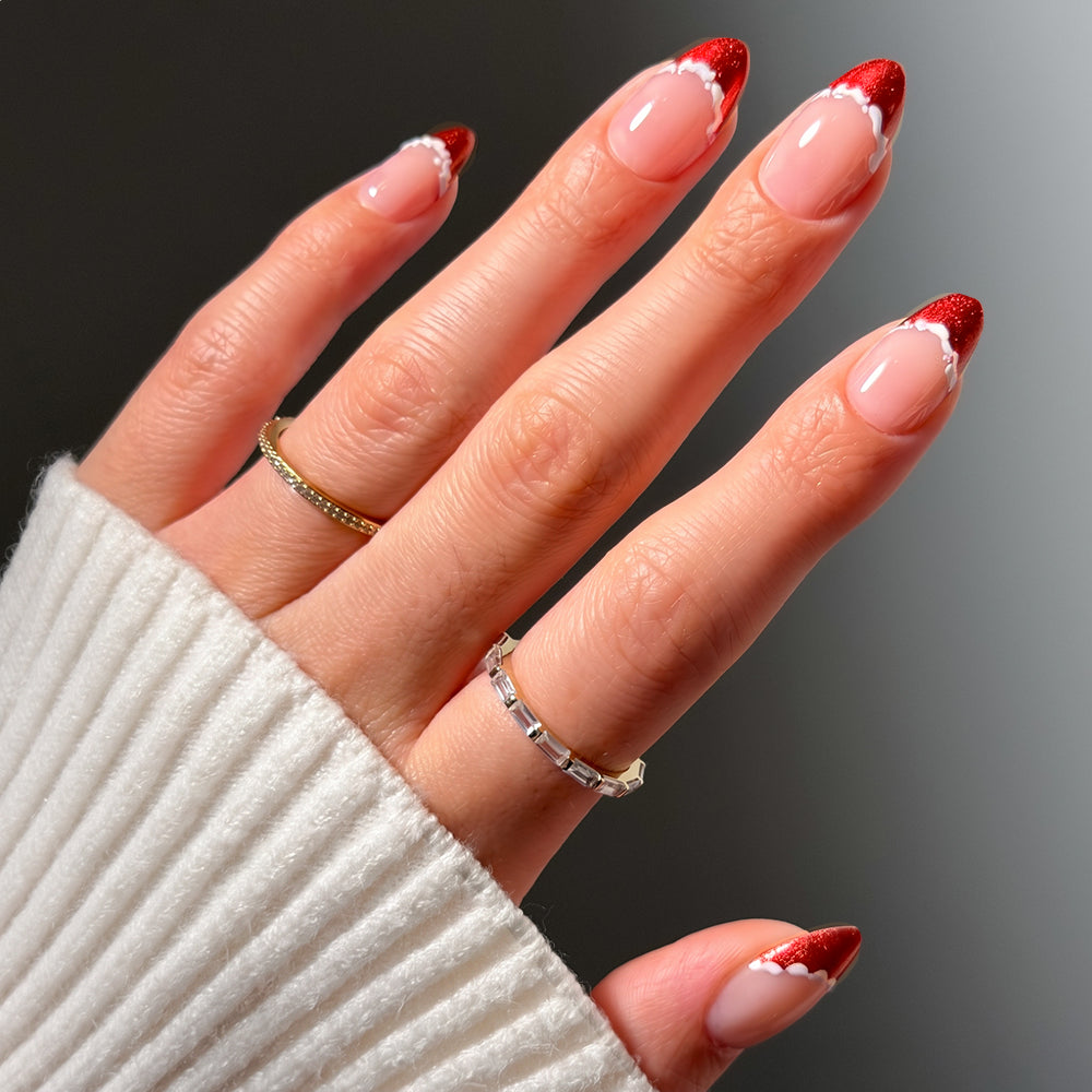 Short almond Snow Kissed press-on nails by glamnetic featuring a nude base with sparkly red French tips outlined in a delicate white scalloped border.