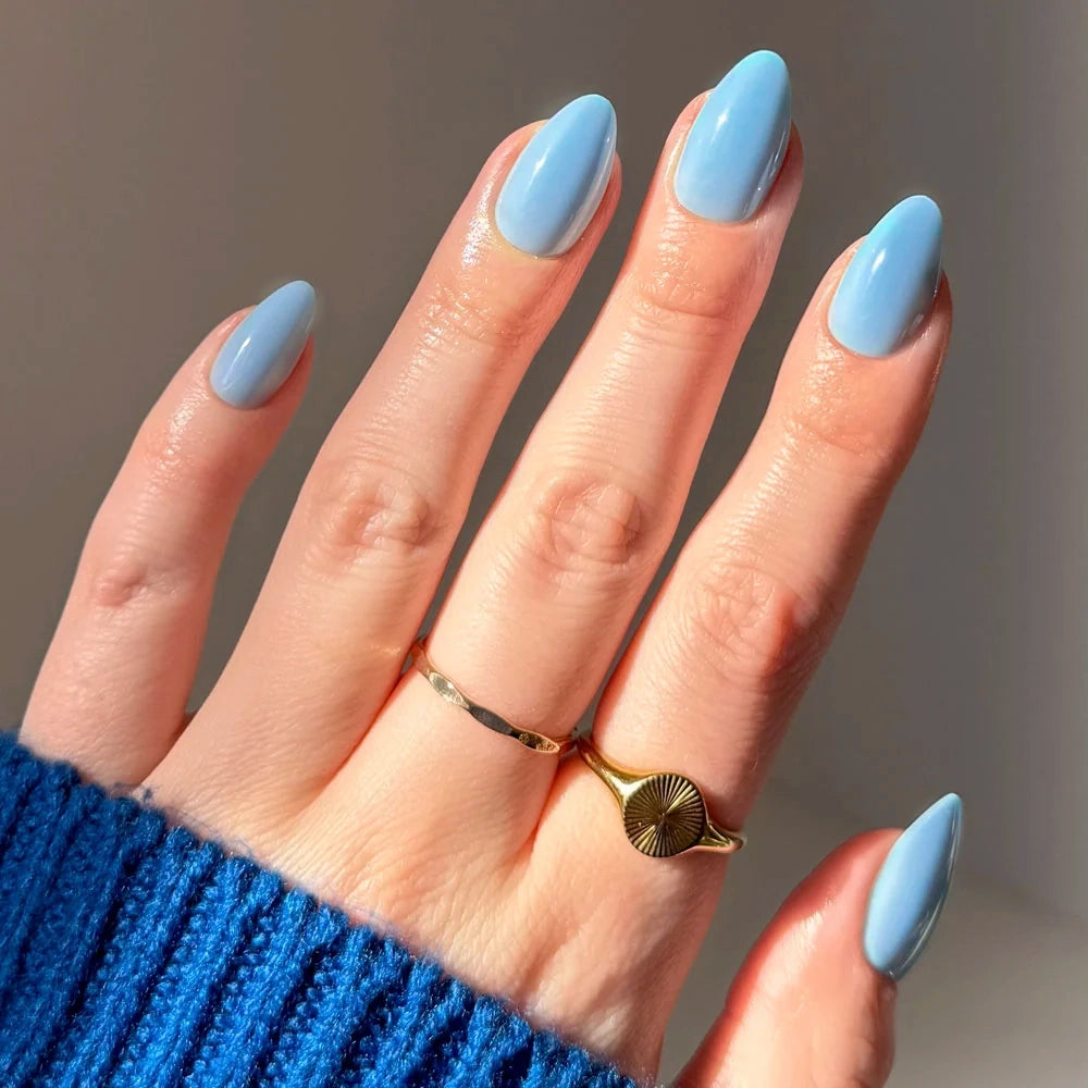 Hand with short almond Glamnetic Soft Blue press-on nails, wearing two gold rings.